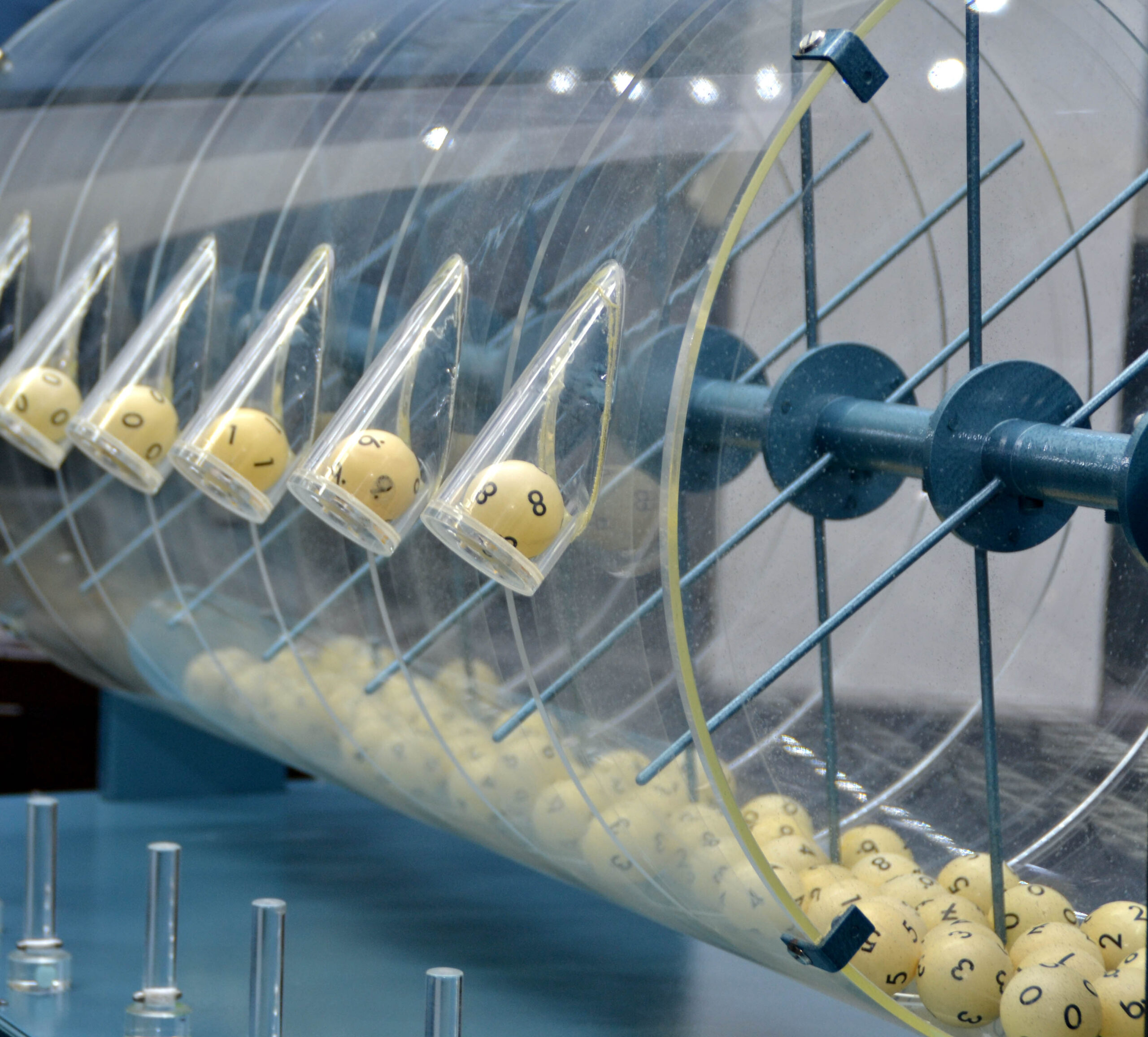 weiße Kugeln mit schwarzem Zahlen-Aufdruck in einer großen durchsichtigen Plastiktrommel. Links im Bild sind in einzelnen Röhren die Kugeln mit den Gewinnzahlen zu sehen.