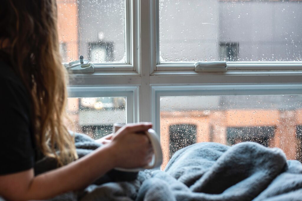 Aktivitäten für Zuhause bei Regenwetter, Frau sitzt vor einem Fenster