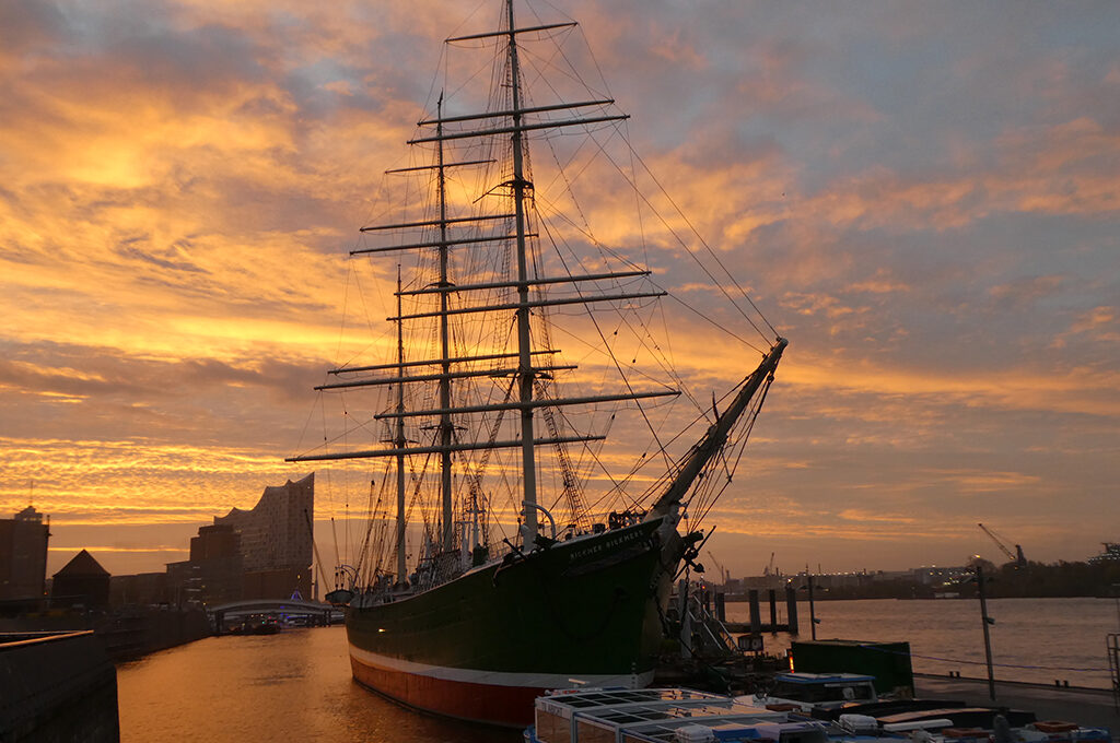 das Segelschiff Rickmer Rickmers liegt am Sonnenuntergang an den Landungsgrücken. Im Hintergrund ist die Elbphilharmonie zu sehen.