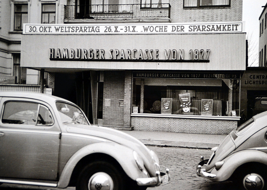 Weltspartag - Weltsparwochen - Kinderkonto: Schwarz-Weiß-Foto von 1959 zeigt alte Haspa-Filiale mit Hinweis auf den weltspartag und die Woche der Sparsamkeit. Vor dem Gebäude stehen zwei alte VW Käfer.