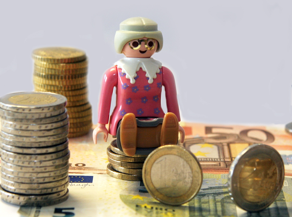 Frauen Finanzen: Eine Playmobil-Omi sitzt auf einem kleinen Stapel Euro-Münzen. Der "Fußboden" sind Euro-Scheine. Neben der Frau stehen einzelne und gestapelte euro-Münzen.