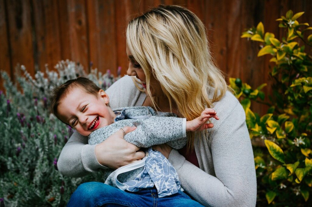 Sparen für Kinder: Blonde Frau vor Garten hält lachendes Kind im Arm