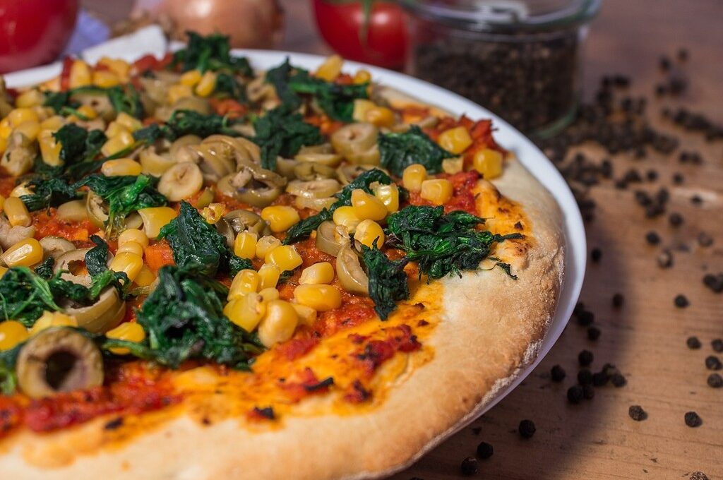 Vegane Pizza in Hamburg: Pizza mit Spinat, Pilzen und Mais auf Holztisch; diverse Zutaten und Pfeffer im Hintergrund