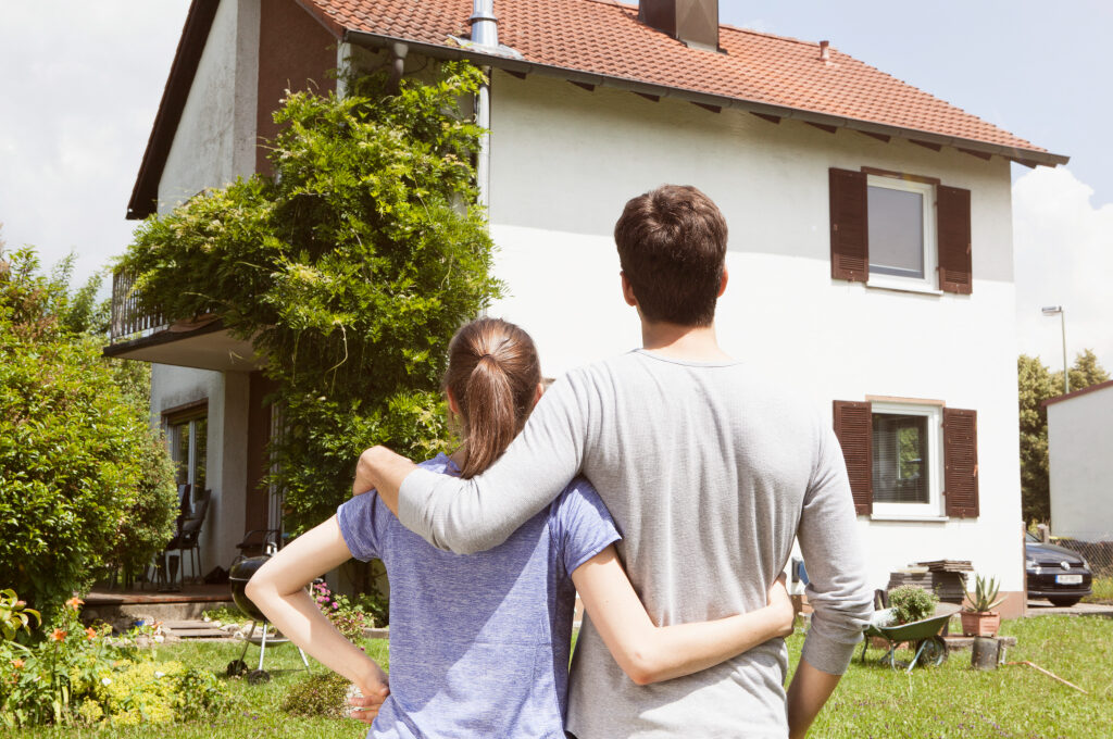 Haus versichern, was braucht man: Paar von hinten zu sehen, steht vor Haus, Arm in Arm
