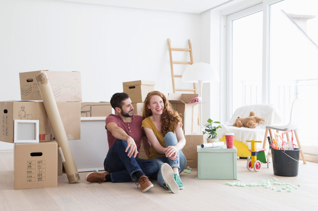 Erste gemeinsame Wohnung, zusammenziehen: Junges Paar sitzt zwischen Umzugskartons und freut sich